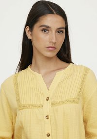 Chemisier jaune à boutons, fabriqué dans un tissu texturé, présentant des plis verticaux et des détails en dentelle le long de l'encolure, avec des boutons ronds.