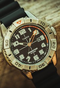 Montre Timex Expedition avec cadran noir, chiffres blancs, aiguille des heures flèche orange, lunette argentée marquée de chiffres et bracelet en caoutchouc noir.