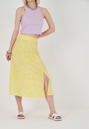 Maxikleid mit gelbem Blumenmuster und einem seitlichen Schlitz, kombiniert mit einem lavendelfarbenen, kurz geschnittenen Tanktop und weißen Rutschsandalen.
