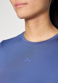 Blauwe sportshirt met een ronde halslijn en korte mouwen. Beschikt over een subtiel logo in variërende tinten blauw op de borst. Gladde textuur.