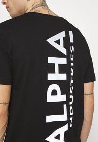 Svart bomull T-skjorte med vertikal hvit trykk av "ALPHA INDUSTRIES" på baksiden. Korte ermer og glatt tekstur.