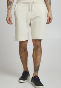 Cremfarbene Sweatshorts mit einem elastischen Bund, Kordelzug, Seitentaschen und umgeschlagenen Säumen. Getragen mit schwarzen Sneakern.