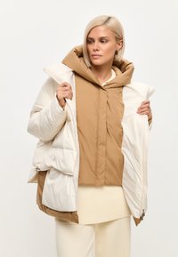 Pustesjakke med en beige yderflade og en cremefarvet inderside, stor hætte, quiltet design og lynlåslommer. Inderlaget er brunt.