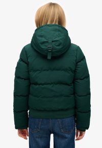 Zielona kurtka puchowa z kapturem, z logo na ramieniu, elastycznymi mankietami i ściągaczem z tyłu do regulacji. Widoczna tkanina dżinsowa.