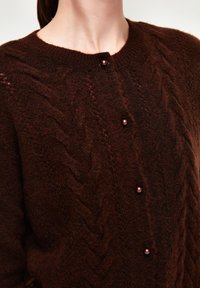 Cardigan en tricot marron avec des motifs en câble, décolleté rond et boutons en forme de perles sur le devant. Texture douce et épaisseur moyenne.