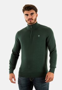 Maglione verde scuro con zip al collo, realizzato in un tessuto liscio ed elasticizzato, con colletto e polsini a coste, dotato di un piccolo logo sul petto.
