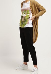 Un long cardigan en tricot ajouré doré sur un t-shirt graphique blanc arborant un design de cactus vert. Un pantalon noir ajusté et des baskets blanches complètent l'ensemble.