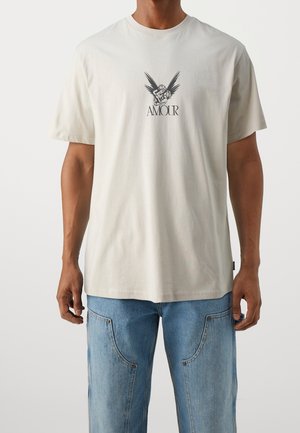 Homme portant un t-shirt beige clair avec un graphisme noir d'un ange enlaçant un enfant et le mot « AMOUR » en dessous, associé à un jean bleu.