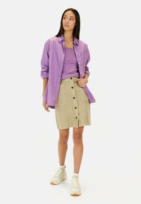 Chemise cintrée lilas au-dessus d'un débardeur lilas, associée à une jupe beige à boutons. Des baskets blanches avec des semelles en gomme complètent l'ensemble.