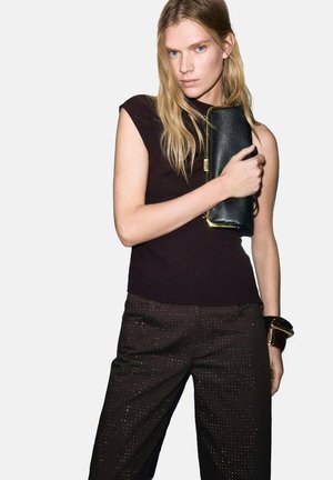 Zwarte leren clutch met gouden sierbeslag, gecombineerd met een nauwsluitend donker topje en bruin, patroonrijke broek met subtiele glanzende accenten.