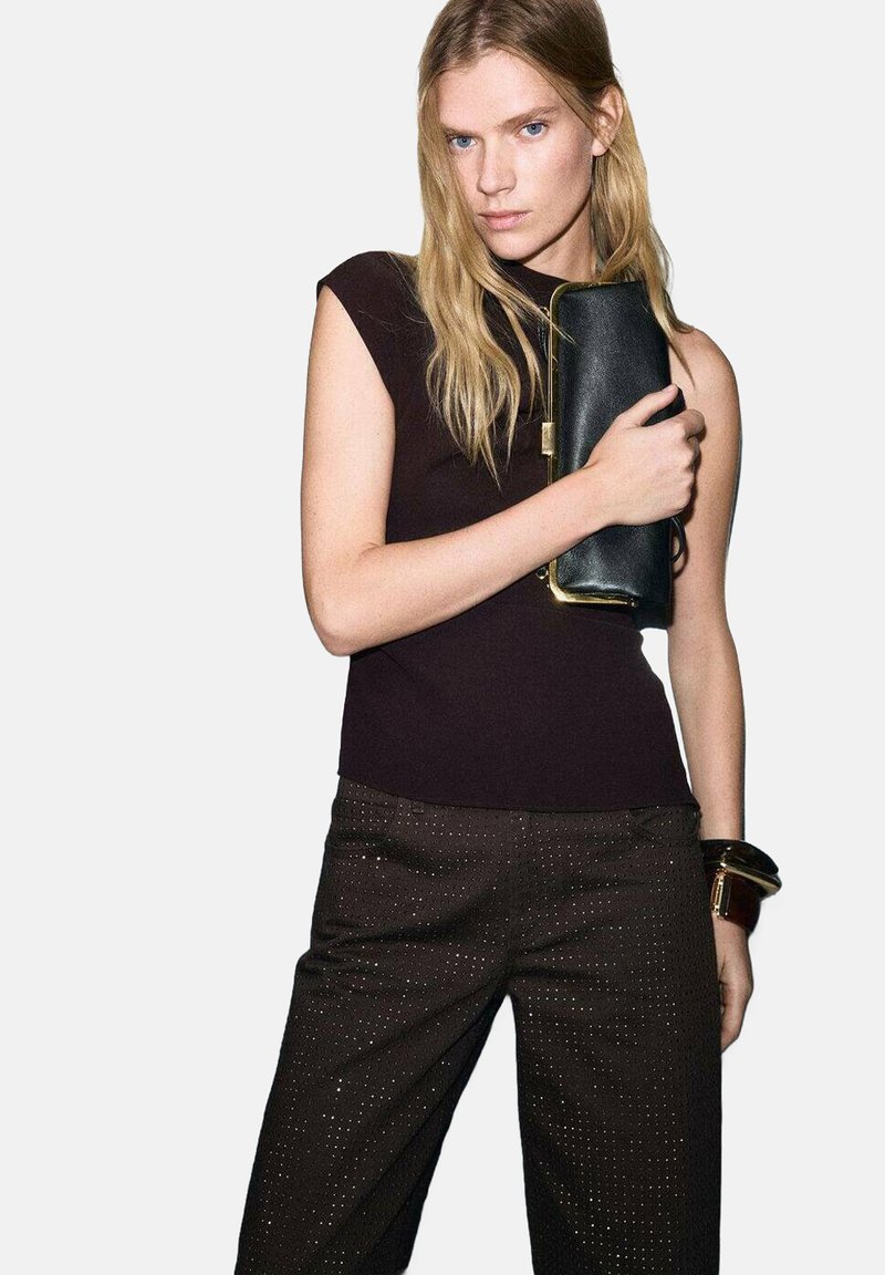 Zwarte leren clutch met gouden sierbeslag, gecombineerd met een nauwsluitend donker topje en bruin, patroonrijke broek met subtiele glanzende accenten.