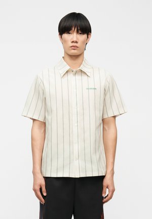 Homme portant une chemise blanche à manches courtes, à boutons, avec de fines rayures verticales et un petit texte vert sur le côté gauche de la poitrine.