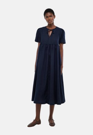 Femme portant une robe midi bleu marine à manches courtes avec un col à nouer, debout les bras détendus, associée à des chaussures plates tissées marron.