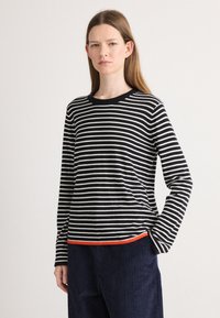 Svart-hvite stripete langermet skjorte med rund hals og oransje kanting på nederste del, kombinert med marineblå cordfløyelsbukser.