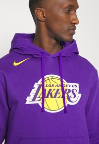 Felpa viola con tasca frontale; presenta "Los Angeles Lakers" e un logo di pallacanestro in giallo e bianco. Logo del marchio sulla spalla.