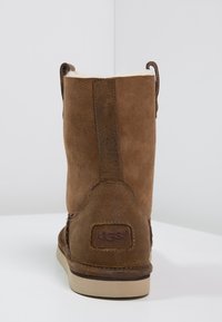 Ruskea nupukkinahkasaapas, jossa on pehmeä sisävuori, pyöreä muoto, ommeltu yksityiskohtia ja kumipohja. "UGG"-logo kantapäällä.