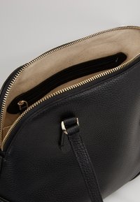 Sort læderhåndtaske med tekstureret overflade og gyldent lynlås. Indvendigt har den en beige foring og en lynlåslomme til organisering.