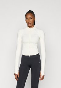 Hvid zip-up atletikjakke med lange ærmer, lavet af et glat strækmateriale. Kombineret med sorte tætsiddende leggings.