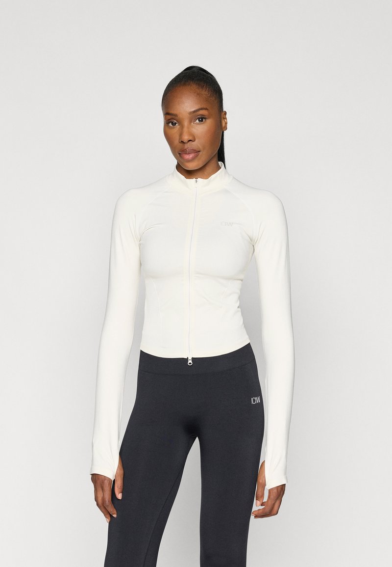 Hvid zip-up atletikjakke med lange ærmer, lavet af et glat strækmateriale. Kombineret med sorte tætsiddende leggings.