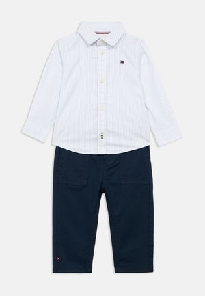 Camisa de manga longa azul claro riscada com fecho de botão e emblema, combinada com calças escuro-marinho com bolsos laterais e um detalhe de logotipo.