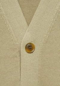 Beige kofta med V-ringning, strukturerat stickmönster och en rund brun knapp vid stängningen. Mjuk kvalitet och avslappnad stil.