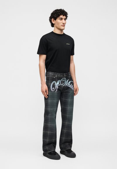 Άνδρας που στέκεται φορώντας μαύρο μπλουζάκι με μικρό λογότυπο και σκούρο καρό παντελόνι με μεγάλο κείμενο "Off-White" στο μπροστινό μέρος.
