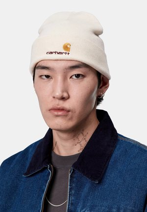 Cremfarbene Mütze mit gesticktem Logo und Akzent, weicher Rippstrickstruktur. Das Modell trägt eine Jeansjacke mit dunklem Cordkragen und einem grauen Hemd.