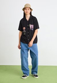 Chemise noire à manches courtes avec des patches brodés colorés, jeans baggy bleus, chaussures noires avec des accents floraux et un chapeau seau beige.