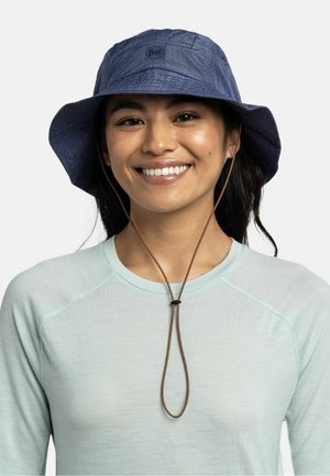 Mujer sonriente con camiseta azul claro y sombrero tipo bucket azul marino con correa ajustable para la barbilla sobre un fondo liso.
