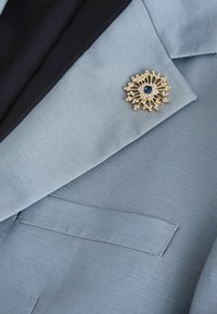 Broche en or et diamants avec une pierre bleue centrale, épinglée sur le revers d'un blazer bleu clair sur un fond en tissu sombre.