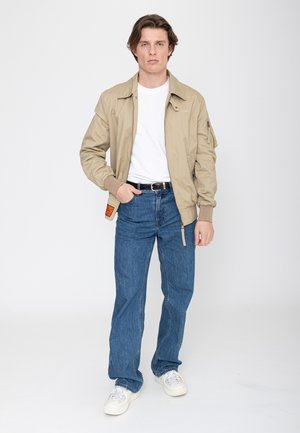 Junger Mann steht und trägt eine beige Jacke, weißes T-Shirt, blaue Jeans, schwarzen Gürtel und weiße Sneakers vor einem neutralen Hintergrund.