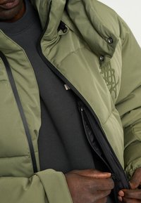 Grøn polstret jakke med lynlås foran og sorte detaljer, med en høj krave og synlige syninger. Underneath er en mørk sweatshirt.