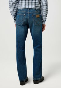 Jean en denim bleu avec une jambe droite, décoloration plus claire au niveau des cuisses, couture contrastante, et deux poches arrière avec un design en zigzag.