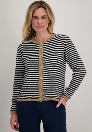 Blonde vrouw die een zwart-wit gestreepte cardigan draagt met knopen, met een beige bies en donkerblauwe broek, staand met één hand in haar zak.
