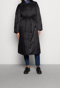 Černý zateplený kabát s límečkem, páskem v pase, dlouhými rukávy a bočními kapsami, nošený přes modré džíny a černé boty.