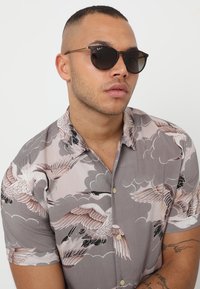 Camisa de manga curta com um fundo cinza, apresentando padrões de grandes aves e nuvens em rosa suave e preto, complementada por óculos de sol arredondados.