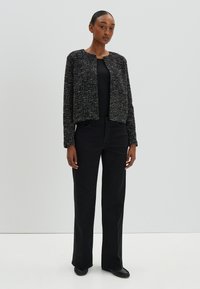 Veste noire cropped avec un tissu texturé, col rond, fermeture à crochet à l'avant, portée avec un pantalon noir taille haute et des chaussures plates.