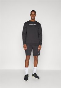 Black sweatshirt with white "HYROX" logo, paired with black shorts. Textured fabric, ribbed cuffs, and black athletic shoes with white accents.