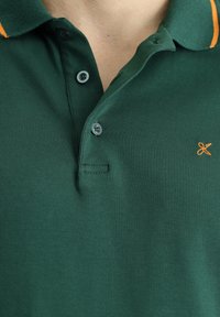 Polo shirt verde scuro con colletto a punta, caratterizzato da una striscia arancione, tre bottoni e un piccolo accento ricamato sul petto.