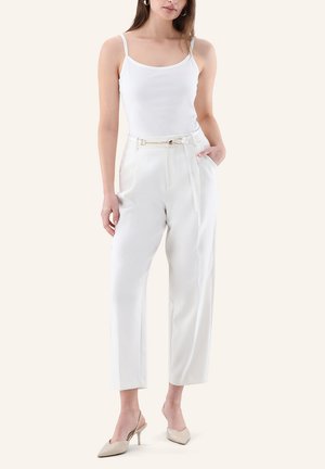 Donna che indossa un top bianco senza maniche, pantaloni bianchi a vita alta con cintura a catena dorata e décolleté beige con punta a contrasto su sfondo neutro.