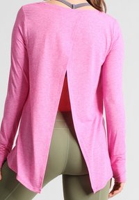 Haut à manches longues en tissu rose clair avec un design à dos partiellement ouvert. Présente un ourlet arrondi et est associé à des leggings verts.