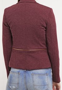 Blazer bordeaux con una vestibilità su misura, caratterizzato da un colletto a pistagna e una cerniera dorata in vita, abbinato a jeans chiari strappati.