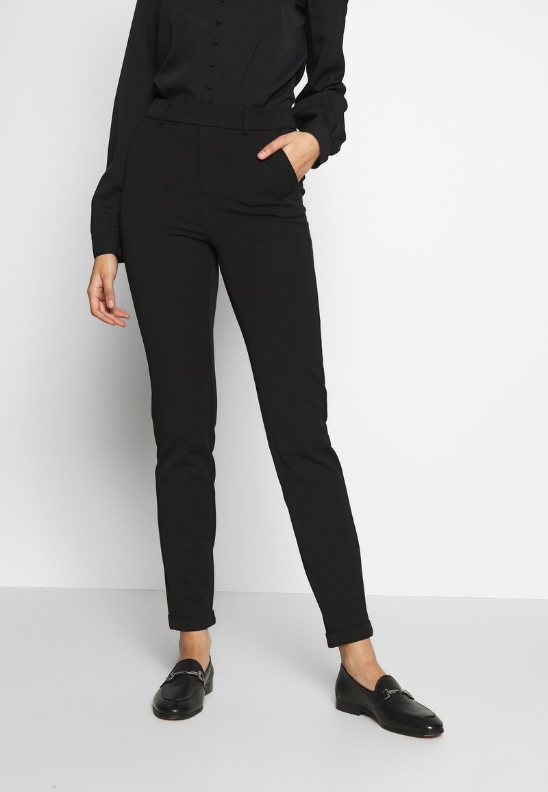 Mustat räätälöidyt housut kapealla istuvuudella, joissa on sileä kangas, sivutaskut ja kääräisty lahje. Yhdistetty mustiin loafer-kenkiin.