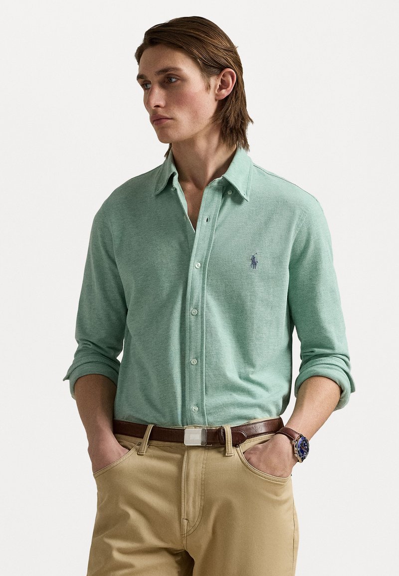 Lichtgroene button-down shirt van zachte stof, met opgerolde mouwen en een klein blauw logo op de borst, gecombineerd met khaki broek en een bruine riem.