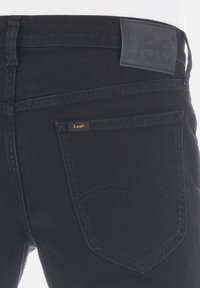 Jeans de mezclilla negra con una textura suave, que presentan una parche de cuero de la marca Lee® en la cintura y una pequeña etiqueta en el bolsillo trasero.