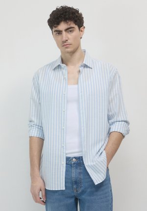 Camicia button-up a righe blu chiaro e bianche, maniche lunghe arrotolate fino ai gomiti, tessuto di cotone, indossata sopra una canottiera bianca. Vestibilità casual.
