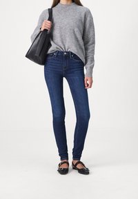 Jeans oscuros ajustados de talle alto, combinados con un suéter de punto gris claro y zapatos planos negros. Se lleva un bolso tote negro.