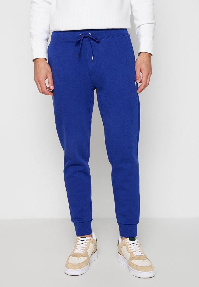 Niebieskie bawełniane spodnie dresowe z elastycznym paskiem w talii, slim fit, bocznymi kieszeniami i ściągaczami przy kostkach; połączone z beżowymi trampkami.