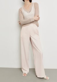 Beige offenes Strickoberteil mit V-Ausschnitt und langen Ärmeln, kombiniert mit hellrosa weit geschnittenen Hosen und neutralen Sandalen mit Absatz. Glatte Textur.