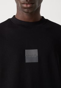 Homme portant un sweat-shirt noir à col rond avec un patch carré portant le logo "HUGO" centré sur la poitrine.